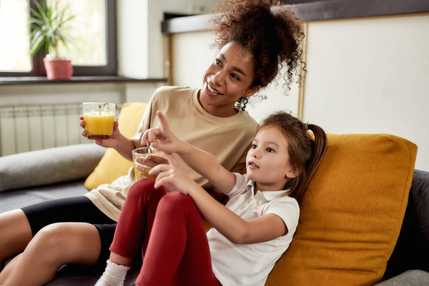 Spending time together. African american woman baby sitter entertaining caucasian cute little girl. They are sitting on the couch, drinking juice while watching TV - Photo, Image