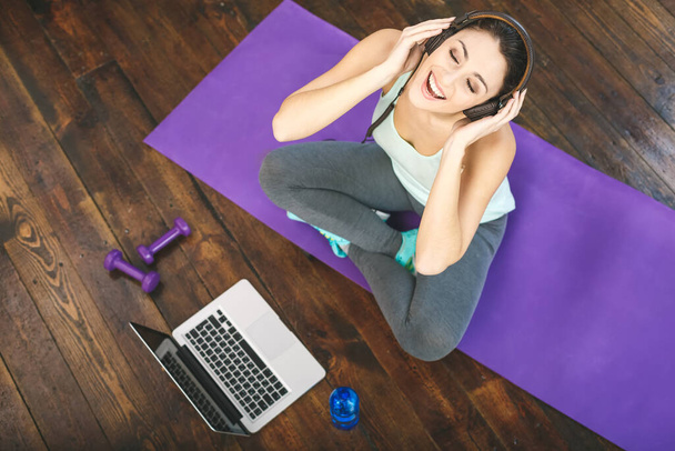 Cheerful beautiful young fitness woman sitting on mat at home and listening to music with headphones. Relax after work-out. Sport concept on wooden dark floor. Top view.  - Foto, imagen
