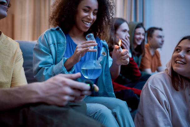 Friends smoking weed at home. Young afro american girl lighting marijuana in the glass bong, relaxing with friends on the sofa at home. Young people playing video games - Foto, afbeelding