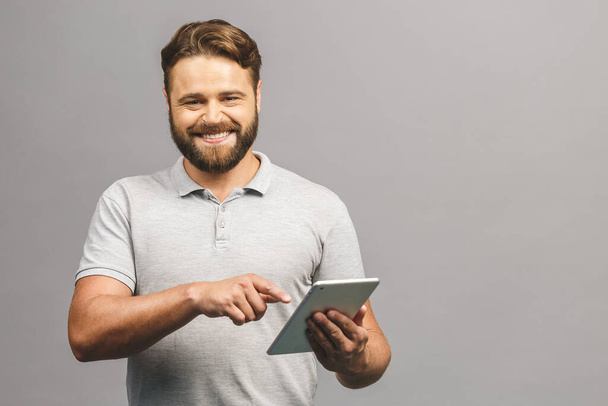 Happy bearded young man in casual standing and using tablet isolated over grey background. - Photo, Image