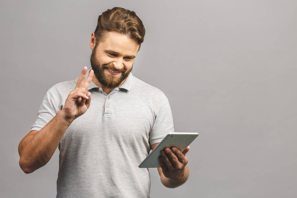 Happy bearded young man in casual standing and using tablet isolated over grey background. - Fotoğraf, Görsel