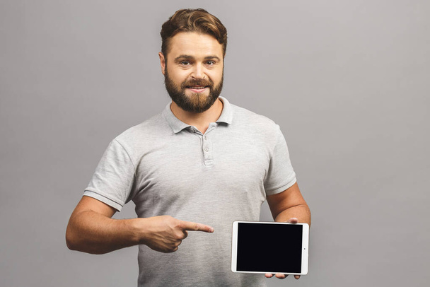 Joyeux jeune homme barbu pointant occasionnellement sur tablette écran vierge isolé sur fond gris
. - Photo, image