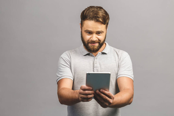 Boldog szakállas fiatalember alkalmi álló és használja tabletta elszigetelt szürke háttér.  - Fotó, kép