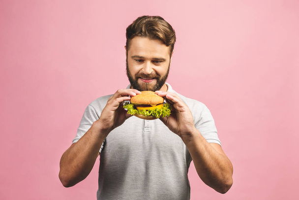 Young man holding a piece of hamburger. Bearded gyu eats fast food. Burger is not helpful food. Very hungry guy. Diet concept. Isolated over pink background. - 写真・画像