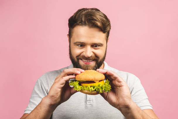 Young man holding a piece of hamburger. Bearded gyu eats fast food. Burger is not helpful food. Very hungry guy. Diet concept. Isolated over pink background. - Photo, Image