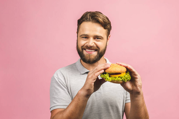 Junger Mann mit einem Stück Hamburger. Bärtiger Gyu isst Fast Food. Burger ist kein hilfreiches Essen. Sehr hungriger Typ. Ernährungskonzept. Isoliert über rosa Hintergrund.  - Foto, Bild