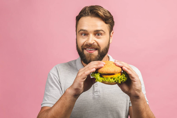 Egy fiatalember egy hamburgert tart a kezében. A szakállas gyu gyorskaját eszik. A hamburger nem segít. Nagyon éhes fickó. Diétás fogalom. Rózsaszín háttérrel elszigetelve.  - Fotó, kép