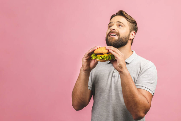 Young man holding a piece of hamburger. Bearded gyu eats fast food. Burger is not helpful food. Very hungry guy. Diet concept. Isolated over pink background.  - Photo, Image