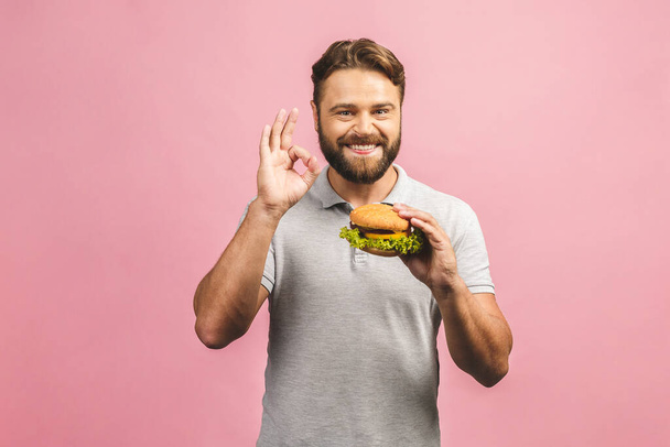 Young man holding a piece of hamburger. Bearded gyu eats fast food. Burger is not helpful food. Very hungry guy. Diet concept. Isolated over pink background. Ok sign. - Photo, Image