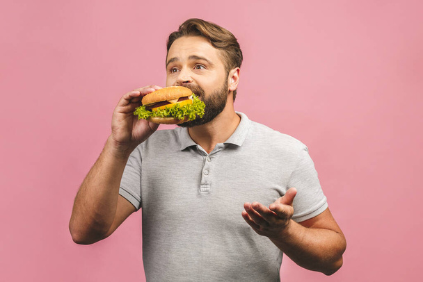 Young man holding a piece of hamburger. Bearded gyu eats fast food. Burger is not helpful food. Very hungry guy. Diet concept. Isolated over pink background. - Foto, immagini