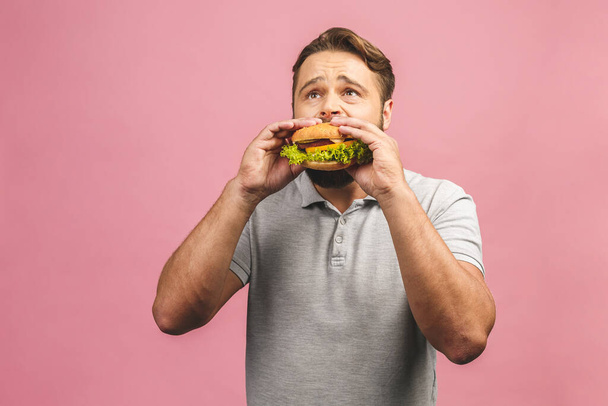 Young man holding a piece of hamburger. Bearded gyu eats fast food. Burger is not helpful food. Very hungry guy. Diet concept. Isolated over pink background. - Foto, Bild