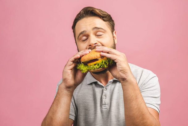 Young man holding a piece of hamburger. Bearded gyu eats fast food. Burger is not helpful food. Very hungry guy. Diet concept. Isolated over pink background. - Fotoğraf, Görsel