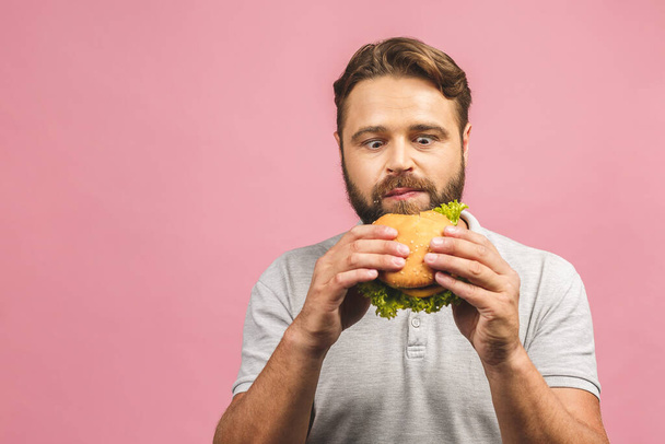 Mladý muž drží kus hamburgeru. Vousatý gyu jí rychlé občerstvení. Burger není užitečné jídlo. Velmi hladový chlap. Dietní koncept. Izolované nad růžovým pozadím. - Fotografie, Obrázek
