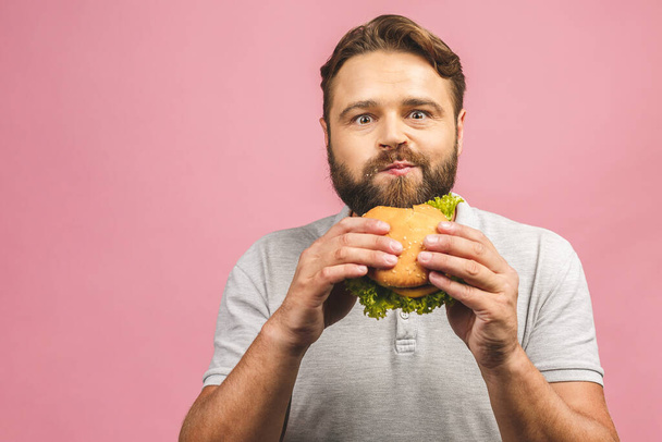 Молодий чоловік тримає шматочок гамбургера. Бородатий гуу їсть фастфуд. Бургери не є корисною їжею. Дуже голодний чоловік. Концепція дієти. Ізольовані над рожевим тлом
. - Фото, зображення