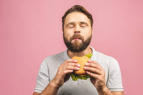 Young man holding a piece of hamburger. Bearded gyu eats fast food. Burger is not helpful food. Very hungry guy. Diet concept. Isolated over pink background. - Photo, Image