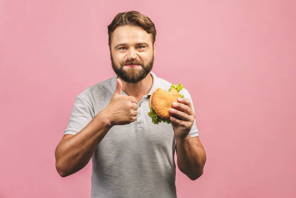 Young man holding a piece of hamburger. Bearded gyu eats fast food. Burger is not helpful food. Very hungry guy. Diet concept. Isolated over pink background. Thumbs up. - Photo, Image