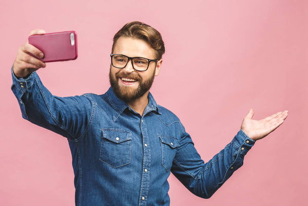 I love selfie! Handsome young man in shirt holding camera and making selfie and smiling while standing against pink background.  - Photo, Image