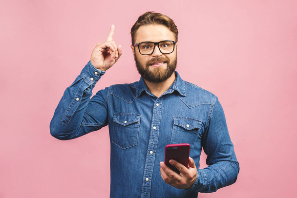 Idea! Young stylish hipster guy dressed in shirt, holds smart phone, reads something attentively, raises index finger, quotes someone words, proves his rightness. People and technology concept. - Photo, Image