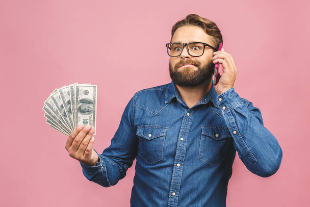 Ευτυχισμένος νικητής! Γενειοφόρος ευτυχισμένος άνθρωπος επιδεικνύοντας χρηματικό έπαθλο του και χρησιμοποιώντας smartphone απομονωμένο σε ροζ φόντο. - Φωτογραφία, εικόνα