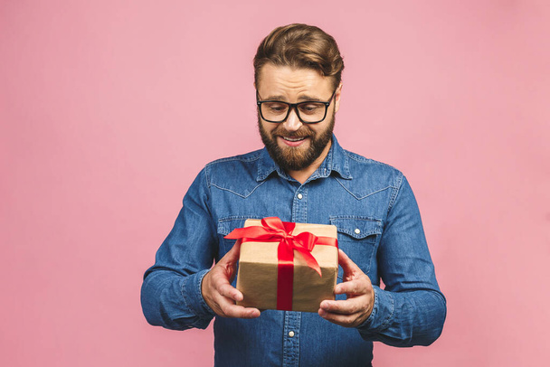 Happy holiday, my congredulations! Portrait of an attractive casual man giving present box and looking at camera isolated over pink background. - Photo, image