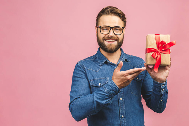 Happy holiday, my congredulations! Portrait of an attractive casual man giving present box and looking at camera isolated over pink background. - Zdjęcie, obraz