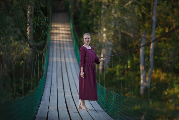 Dívka ve vínově červených šatech kráčí po visutém dřevěném mostě. Usmívající se blondýna - Fotografie, Obrázek