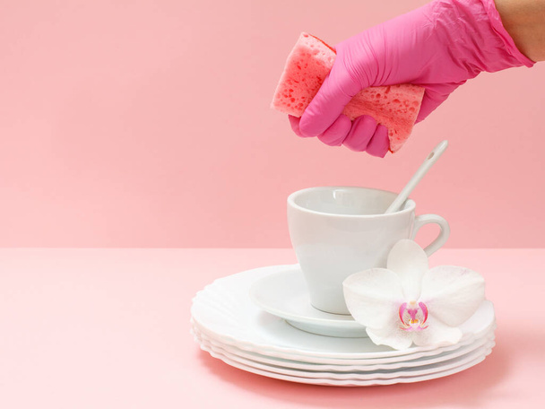 スポンジ、ホワイトプレート、ソーサー、ピンクの背景にカップを持つ硝酸塩使い捨て手袋の女性の手。洗浄・洗浄コンセプト. - 写真・画像
