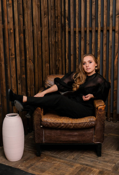 Elegante blonde Dame posiert in schwarzer Bluse mit Schuhen. Modische Frau sitzt auf einem großen Lederstuhl. Holzwand auf dem Hintergrund. - Foto, Bild