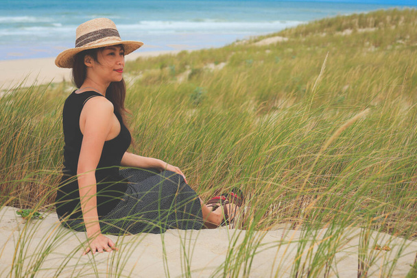 Aasian nainen istuu hiekkadyyneillä, hiekkarannalla ja katsoo kaukaisuuteen - Valokuva, kuva