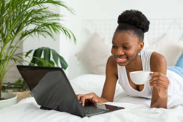 beautiful young african woman lies on bed with laptop - Фото, изображение
