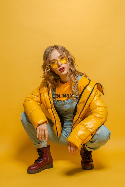 Atractiva mujer modelo de moda en chaqueta de invierno y gafas de sol posando sobre fondo amarillo brillante
 - Foto, imagen
