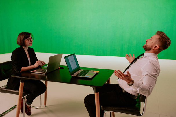 Bonita manjedoura de escritório e empresário têm uma discussão no local de trabalho eles têm dois laptops na frente deles discutindo juntos em um grande estúdio verde
 - Foto, Imagem