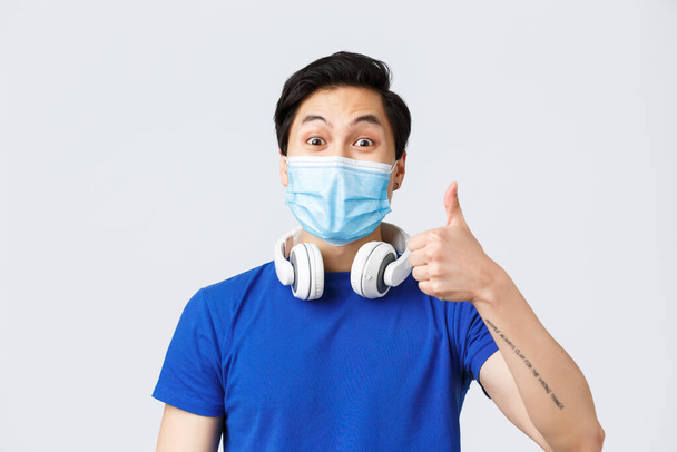 Different emotions, lifestyle and leisure during coronavirus, covid-19 concept. Close-up of upbeat asian guy in medical mask show thumb-up in approval of awesome new song playing in headphones - Photo, Image