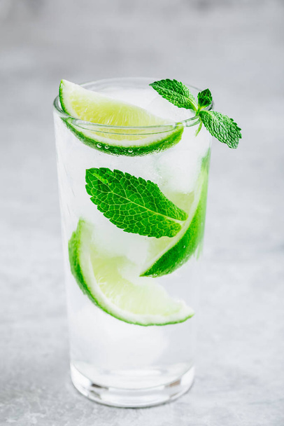 Мохито коктейль со льдом, свежей мятой и лаймом в стекле на сером каменном фоне. Освежающий холодный летний напиток
. - Фото, изображение