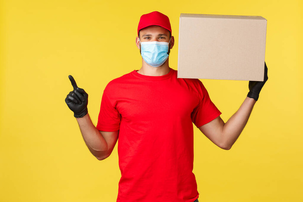 Express delivery during pandemic, covid-19, safe shipping, online shopping concept. Handsome delivery guy, courier holding box with package and pointing upper left corner, wear medical mask - Photo, image