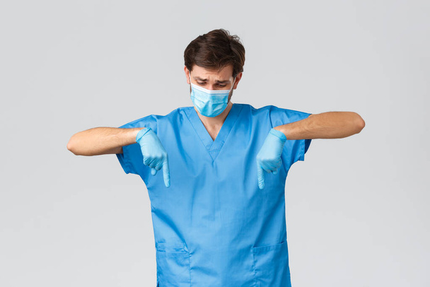 Coronavirus outbreak, healthcare workers fighting disease, hospitals concept. Disappointed doctor showing compassion or sympathy, pointing fingers down, look upset, wear scrubs and gloves - Photo, Image