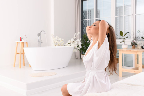 a young woman is sitting on the edge of the bed in a white robe, and a Girl is letting her hair down before taking a bath. Eyes look to the side. - Foto, Imagen