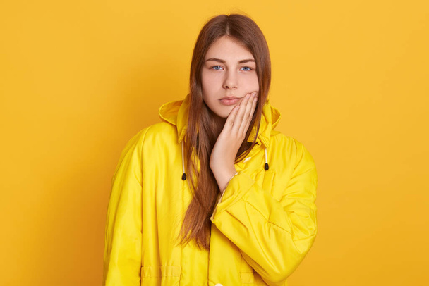 Upset female wearing yellow jacket touching her cheek, suffering from toothache, looks at camera, having medical problems, standing against yellow wall. - Photo, Image