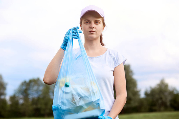 Caucasian young woman picking up trash from meadow. Female cleaning field, collecting litter in garbage bag, wearing t shirt and base ball cap, standing with trees and sky in background. - Photo, Image