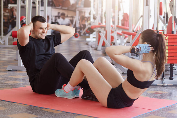 Young attractive Caucasian couple exercising together at gym. Fit man and woman sitting on floor in front of each other and doing abdominal exercises, keeping hands behind heads. - Foto, immagini