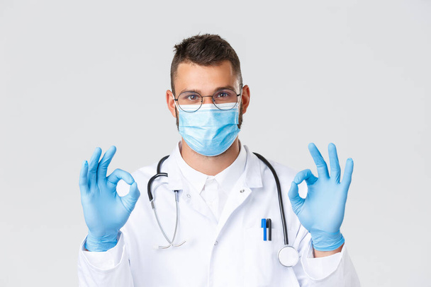 Covid-19 、医療従事者、パンデミック、ウイルスの概念の防止。白いコートと眼鏡で満足ハンサムな医師のクローズアップ,医療マスクの医師は大丈夫サインを示す,プロモーションをお勧めします - 写真・画像