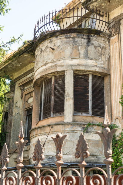Façade d'une vieille maison abandonnée avec des fenêtres en ruine et une terrasse derrière une clôture à motifs rouillés contre un ciel bleu clair
 - Photo, image