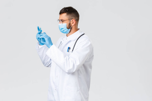 Covid-19, healthcare workers, pandemic, virus concept. Profile shot of serious and focused doctor in white coat, medical mask and gloves, prepare syringe with coronavirus vaccine to give patient shot - Foto, Bild