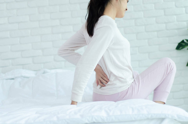Kadınlar sabah kalça ağrısı ile bir odada yatakta beyaz gömlek ve pembe pantolon giymek. Asyalı kadınlar sabah sırt ağrısı var. Sırt ağrısı bir şilte neden olur. Nesnelere odaklanmayın. - Fotoğraf, Görsel