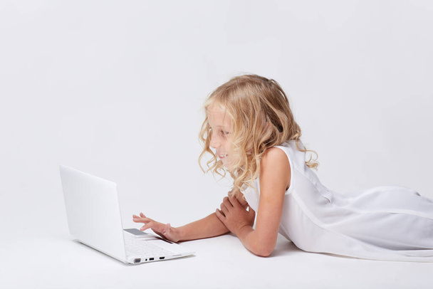 Beautiful and cute blonde little girl plays with white computer notebook laying on floor in bright studio, in white dress. - Photo, image