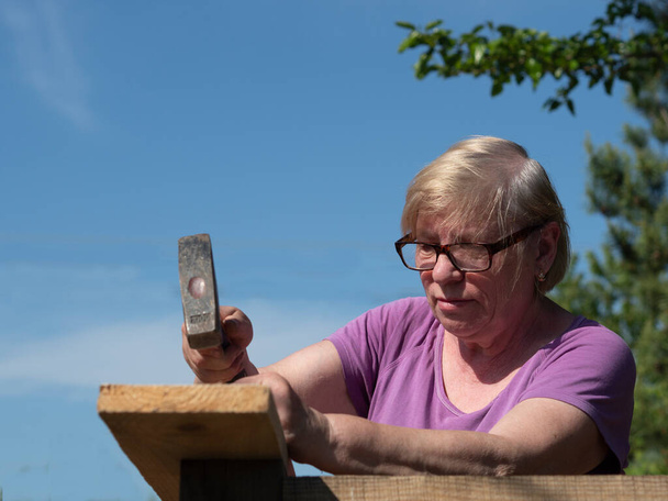 Senior Caucasian woman fastens a board on the roof of a farm building in the garden with a hummer - Valokuva, kuva