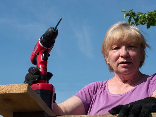 Senior Caucasian woman fastens a board on the roof of a farm building in the garden with a screwdriver - Фото, изображение