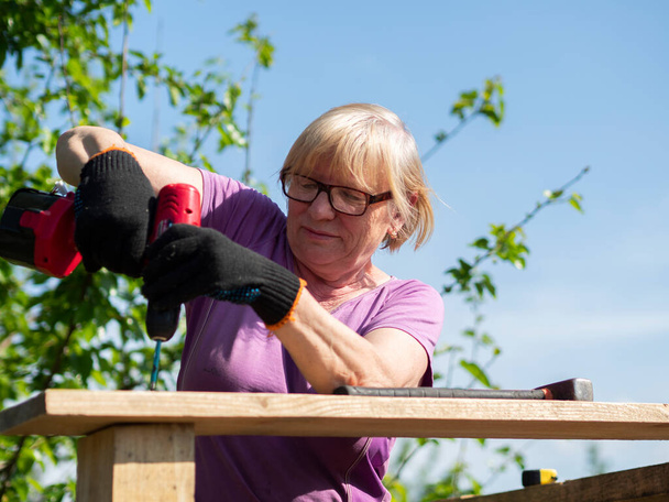Senior Caucasian woman fastens a board on the roof of a farm building in the garden with a screwdriver - Valokuva, kuva