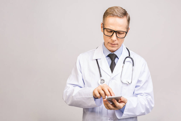 Foto de primer plano del médico senior de pie aislado sobre fondo gris, utilizando el teléfono móvil
. - Foto, Imagen