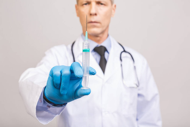 A kék kesztyűs idősebb orvos egy injekcióhoz való orvosi fecskendőt tart szürke alapon elkülönítve..  - Fotó, kép